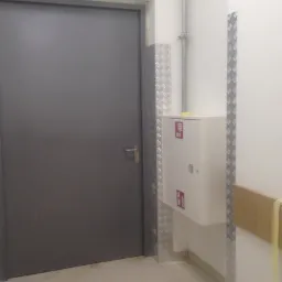 Wnętrze korytarza z szarą, metalową futryną i drzwiami, obudowaną skrzynką hydrantową z oznaczeniami przeciwpożarowymi, zabezpieczonymi ścianami z blachy ryflowanej i rurą instalacyjną.