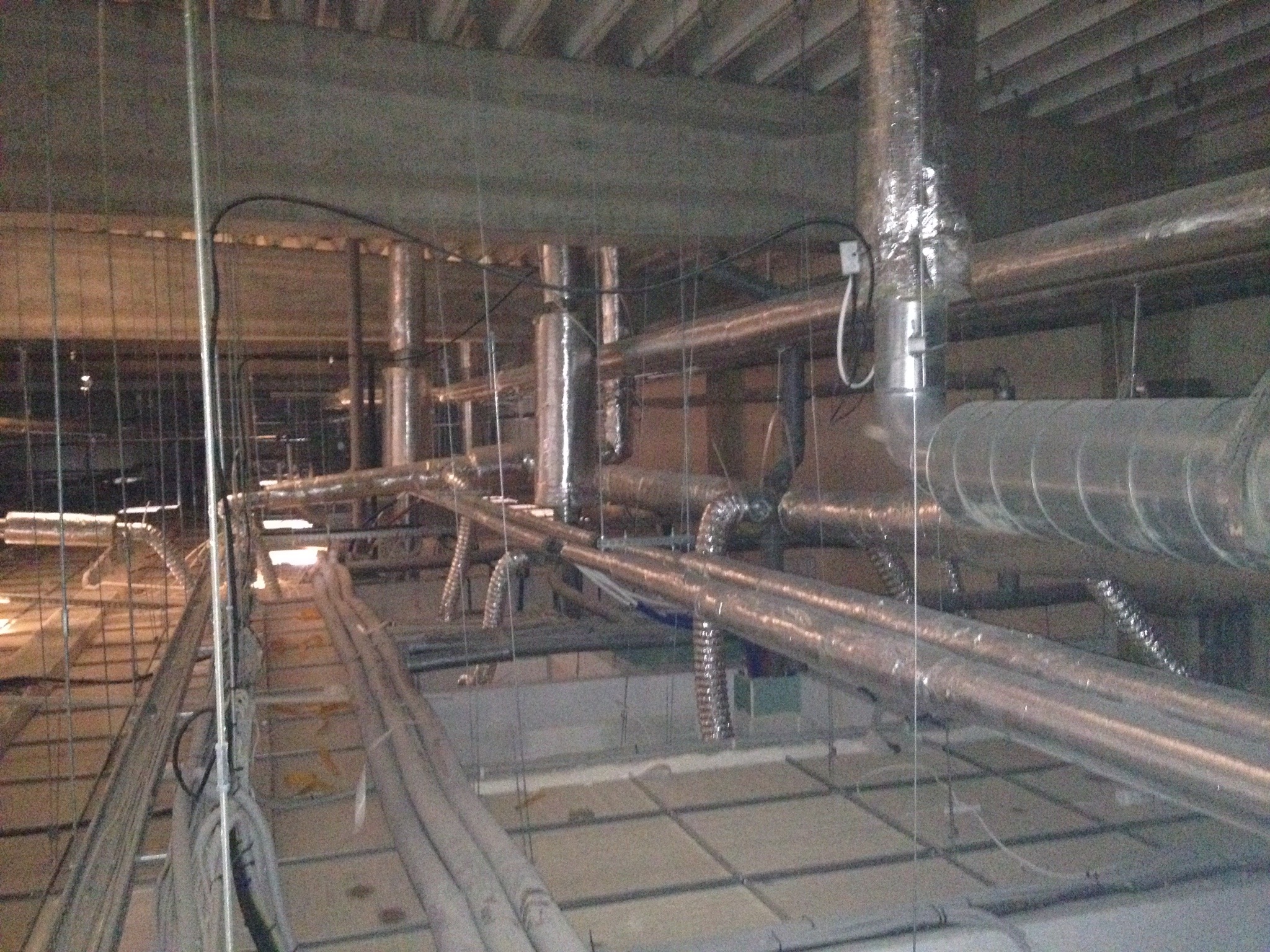 Widok instalacji wentylacyjnych i rur pod sufitem w surowym stanie, z widocznymi przewodami, izolacją i elementami mocującymi.