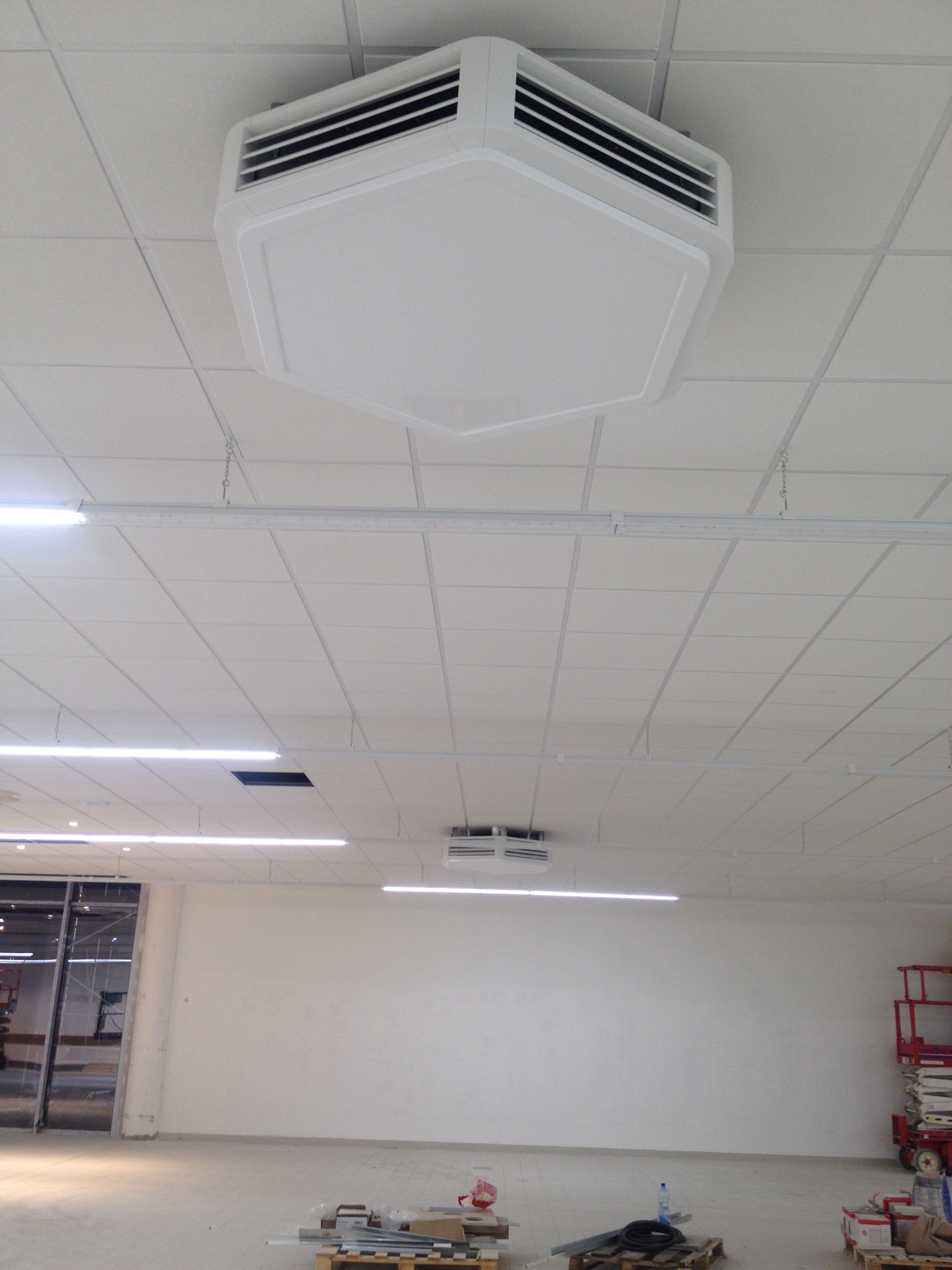 Widok sufitu w pomieszczeniu komercyjnym z zamontowanym, sześciokątnym, białym klimatyzatorem kasetonowym z czarnymi kratkami wentylacyjnymi, oświetleniem LED i podwieszanym sufitem kasetonowym.