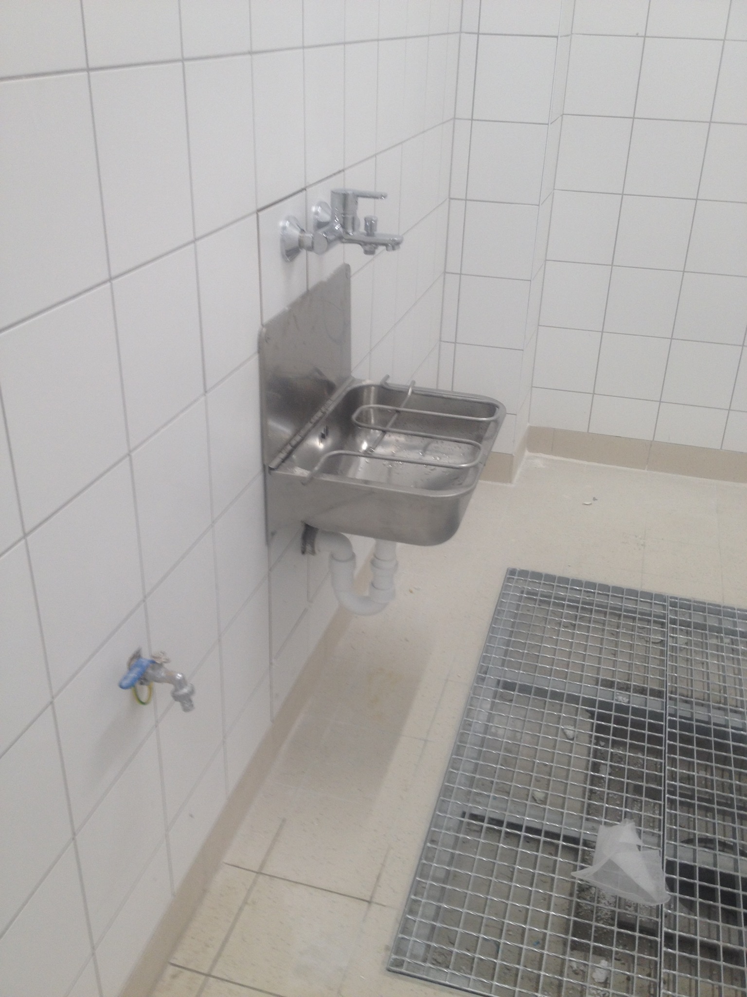 Nierdzewna umywalka przemysłowa z kratką odpływową w pomieszczeniu wyłożonym białymi płytkami.