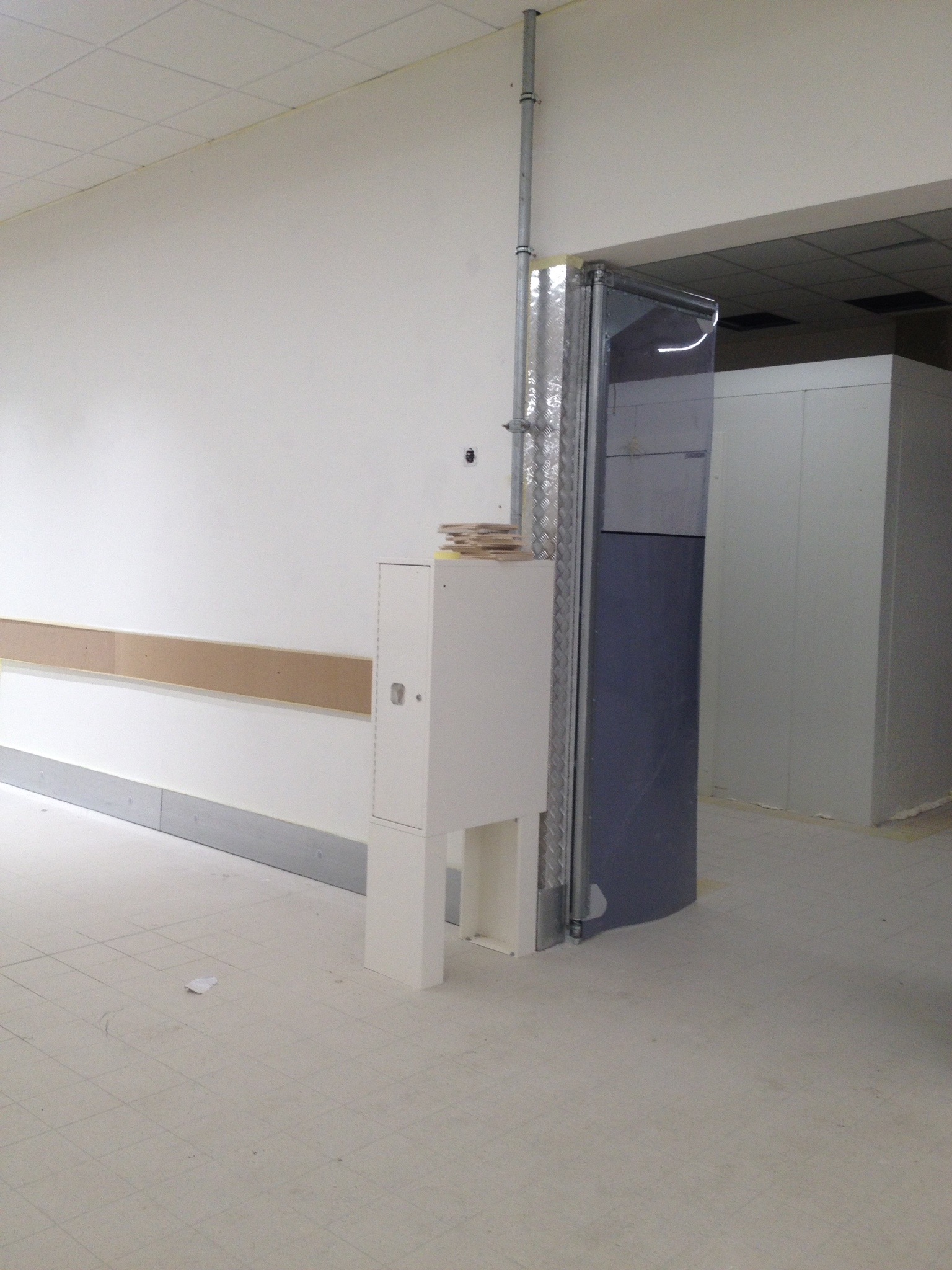 Wnętrze w trakcie remontu: metalowa rura pionowa przy ścianie, biała szafka z dokumentami na górze, częściowo zamontowana listwa przypodłogowa i zabezpieczenie ściany, widoczne wejście do chłodni.