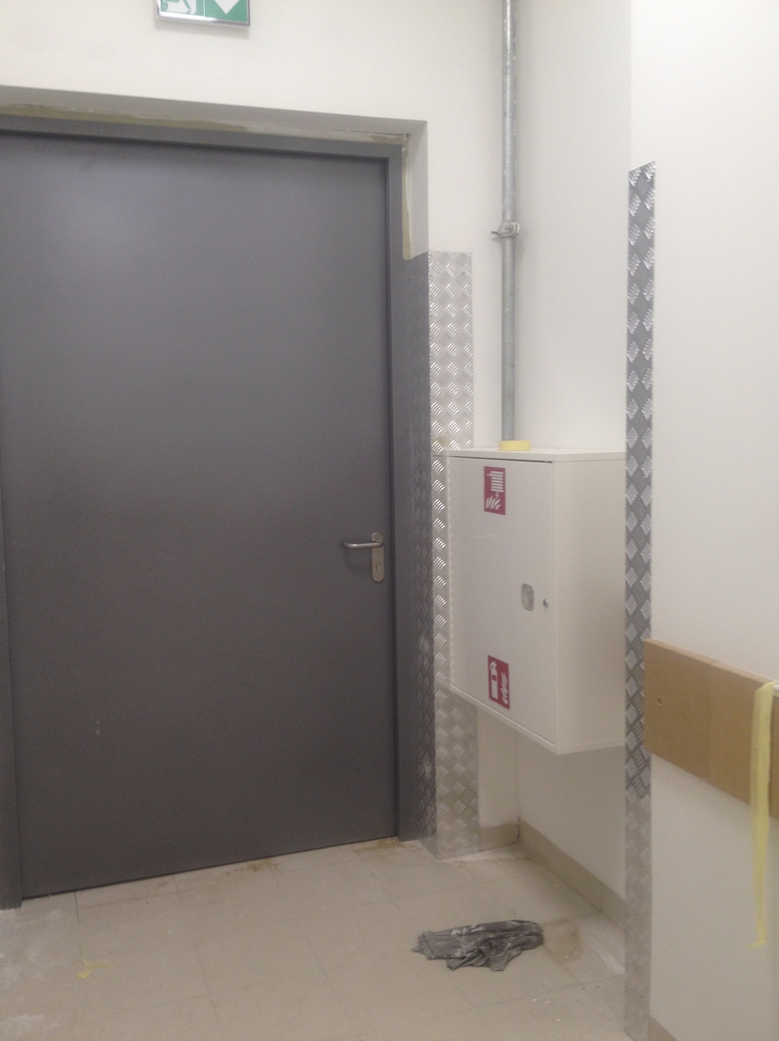 Wnętrze korytarza z szarą, metalową futryną i drzwiami, obudowaną skrzynką hydrantową z oznaczeniami przeciwpożarowymi, zabezpieczonymi ścianami z blachy ryflowanej i rurą instalacyjną.