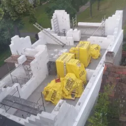 Budowa domu z białych bloczków Ytong, widok z góry, na dachu budynku leżą żółte opakowania z bloczkami, w tle zielone drzewa i dach innego budynku.