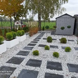 Ogród z ścieżką z kwadratowych płyt na białym żwirze, z kulistymi krzewami i strzyżonymi drzewkami, w tle domek narzędziowy i mały domek dla dzieci.