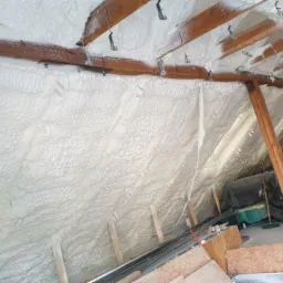 Poddasze ocieplone pianką PUR, widoczne drewniane elementy konstrukcyjne i metalowe uchwyty mocujące, z boku czarna folia zabezpieczająca.