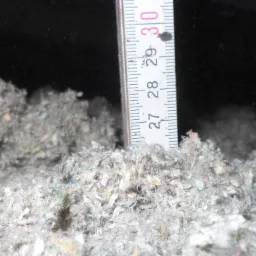 Pomiar grubości warstwy izolacji celulozowej w ciemnym pomieszczeniu, widoczna miarka od 27 do 30 centymetrów.