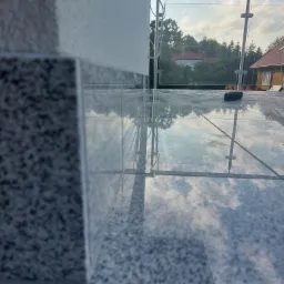 Błyszczące, szare płytki na balkonie z szklaną balustradą, odbijające błękitne niebo i chmury, widok z niskiego kąta.