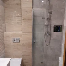 Nowoczesna łazienka z kabiną prysznicową typu walk-in, wykończona płytkami imitującymi drewno i kamień, z wbudowaną półką na kosmetyki i ukrytym systemem spłukiwania toalety.