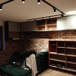 BAJBUD - Wnętrze pokoju z ceglaną ścianą, zabudową meblową pod sufitem, oświetleniem szynowym i zieloną sofą z rzeczami osobistymi.