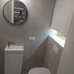 BAJBUD - Nowoczesna łazienka w odcieniach szarości z białym wyposażeniem i złotą armaturą, okrągłe lustro z odbiciem osoby robiącej zdjęcie.