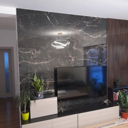 BAJBUD - Salon z czarną ścianą imitującą marmur, drewnianymi lamelami, podświetlanym okrągłym żyrandolem i niską szafką pod telewizorem z roślinami doniczkowymi.
