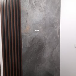 BAJBUD - Szara ściana wykończona płytami imitującymi marmur, sąsiadująca z drewnianą ścianką działową z ciemnymi listwami i białą ścianą, z fragmentem czarnego pufa w dolnej części kadru.