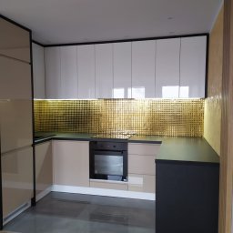 BAJBUD - Nowoczesna kuchnia z błyszczącymi, białymi szafkami górnymi, beżowymi szafkami dolnymi i złotą mozaiką nad blatem. Czarny blat kontrastuje z jasnymi elementami. Podłoga wyłożona szarymi płytkami...