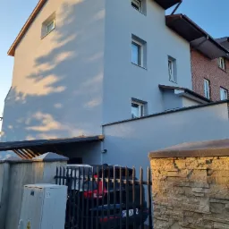 Dwupiętrowy dom z elewacją w odcieniach szarości, widoczny zza metalowego ogrodzenia i kamiennego murku, z zaparkowanym czarnym samochodem pod wiatą.