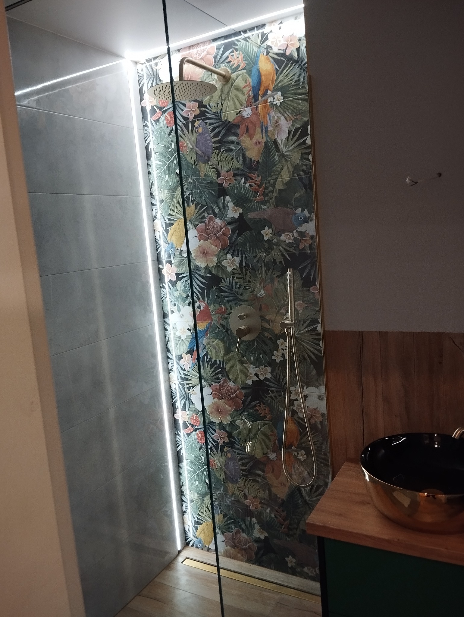 Nowoczesna kabina prysznicowa z szarymi płytkami i ścianą z motywem tropikalnym, oświetlenie LED wzdłuż krawędzi, złota armatura i czarna umywalka na drewnianym blacie.