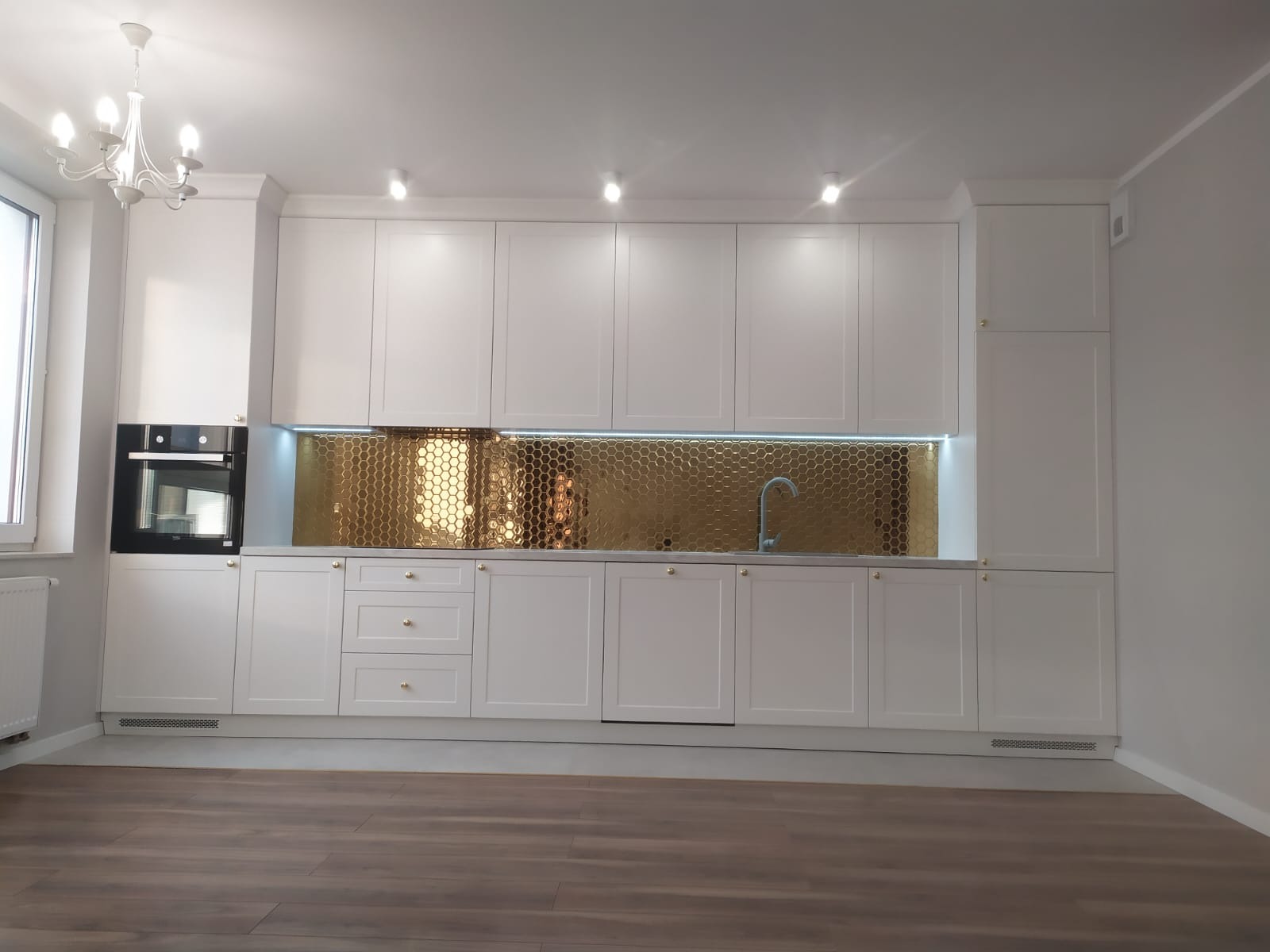 Nowoczesna kuchnia z białymi szafkami i złotą mozaiką heksagonalną nad blatem, oświetlona listwą LED, z drewnianą podłogą i minimalistycznym żyrandolem.