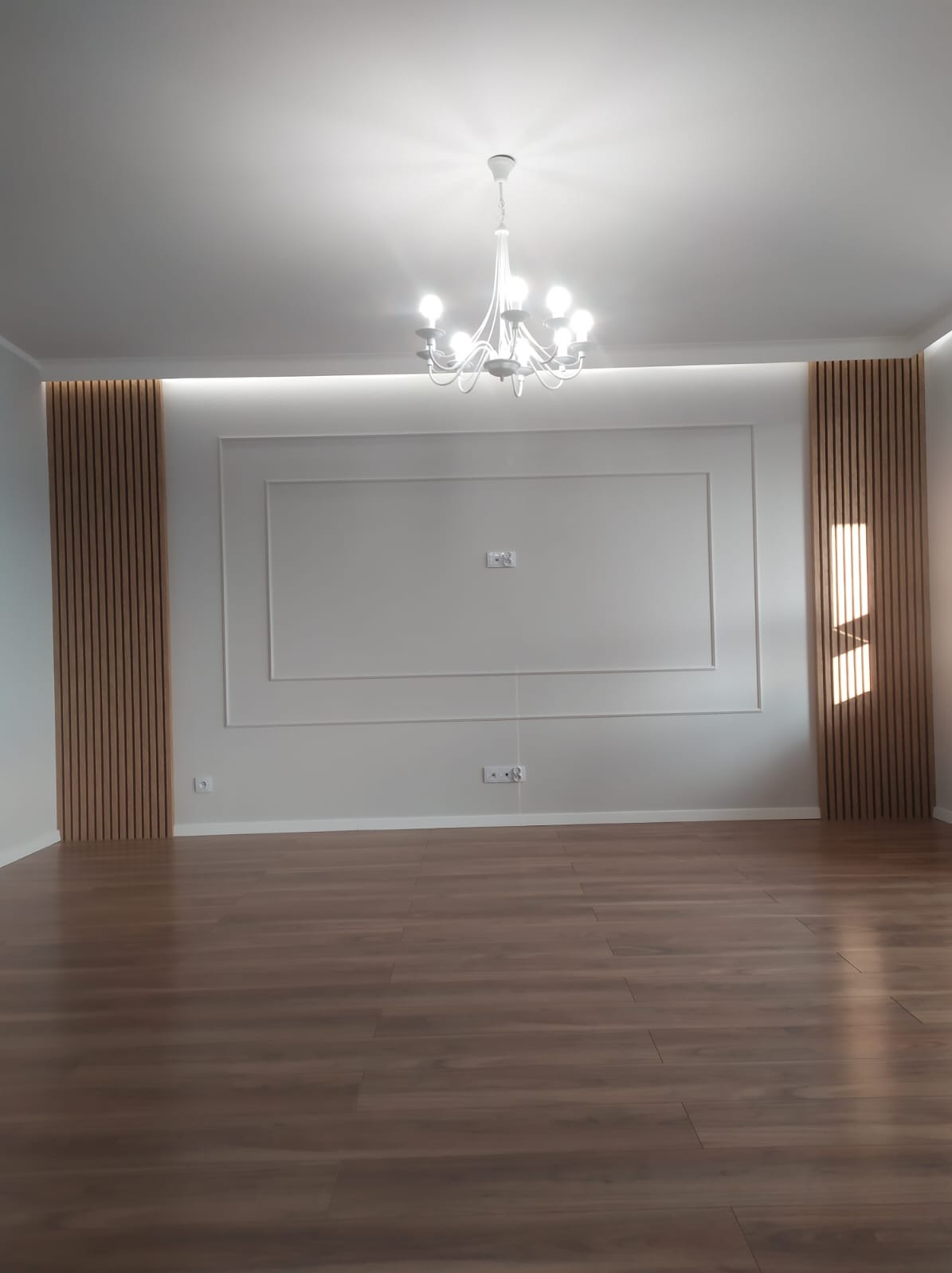 Pusty pokój po remoncie z panelami dekoracyjnymi, sztukaterią i nową podłogą, oświetlony przez biały żyrandol.