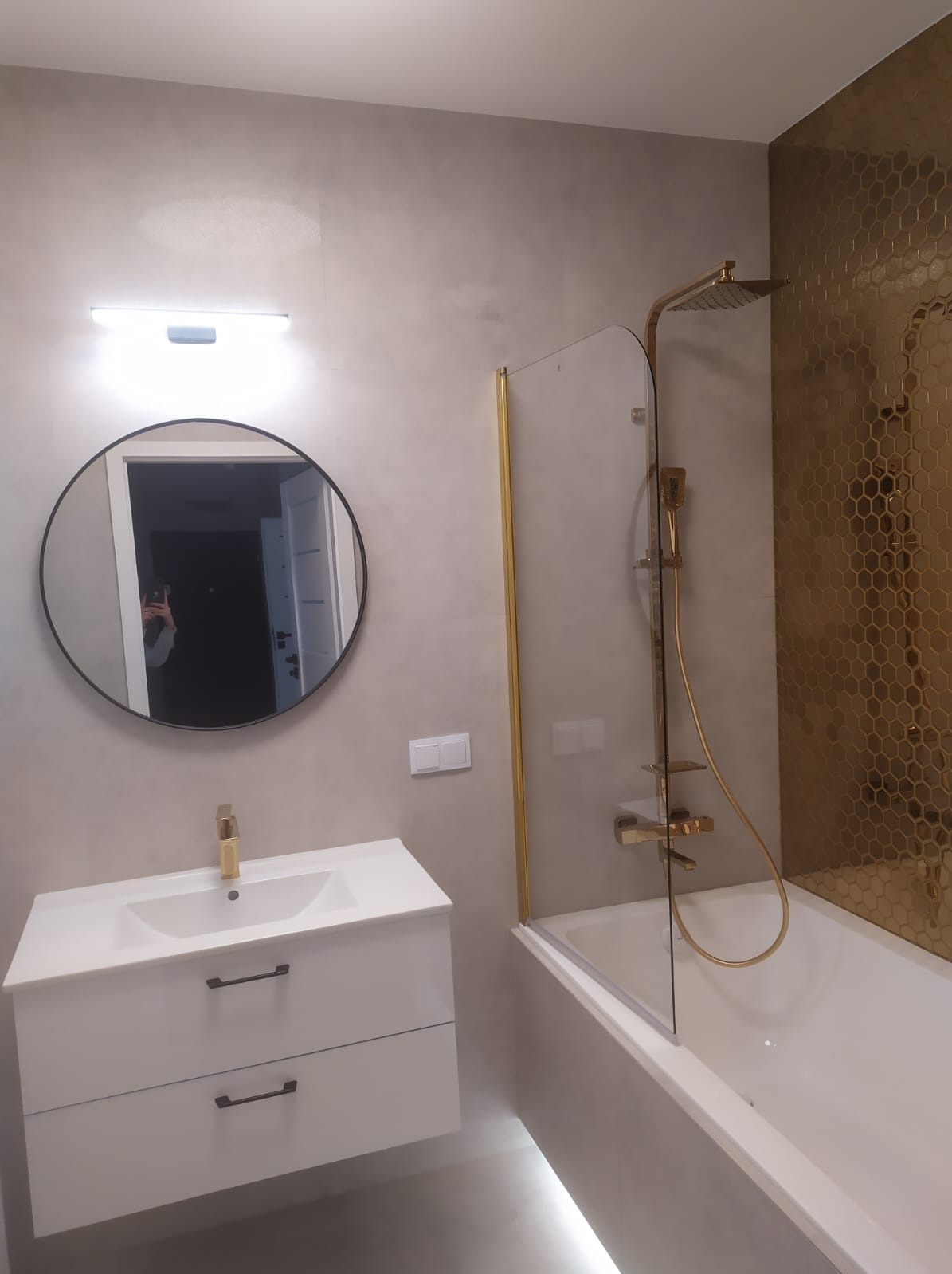 Nowoczesna łazienka z umywalką na szafce, okrągłym lustrem i złotą armaturą oraz ścianą wyłożoną mozaiką w kształcie plastra miodu.