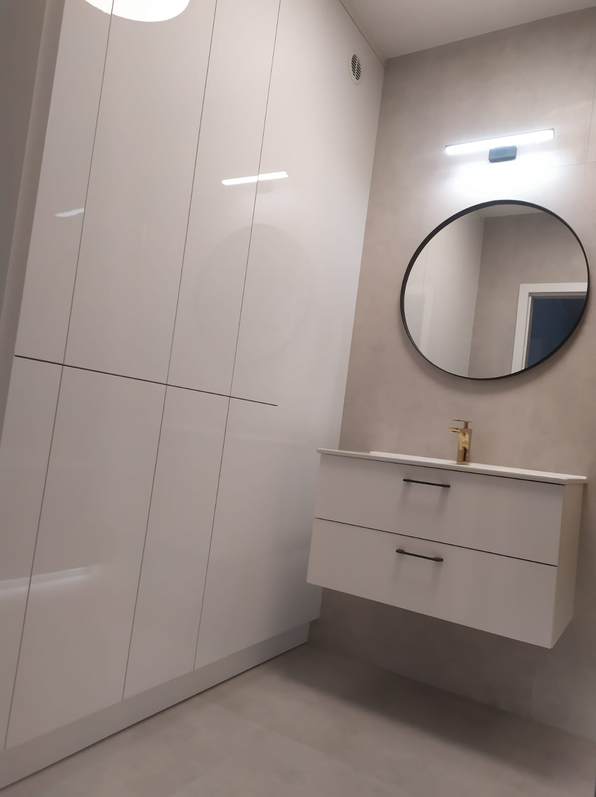 Nowoczesna łazienka z białymi, lakierowanymi szafkami, okrągłym lustrem z czarną ramą i złotą baterią umywalkową podświetlona ledowym światłem.