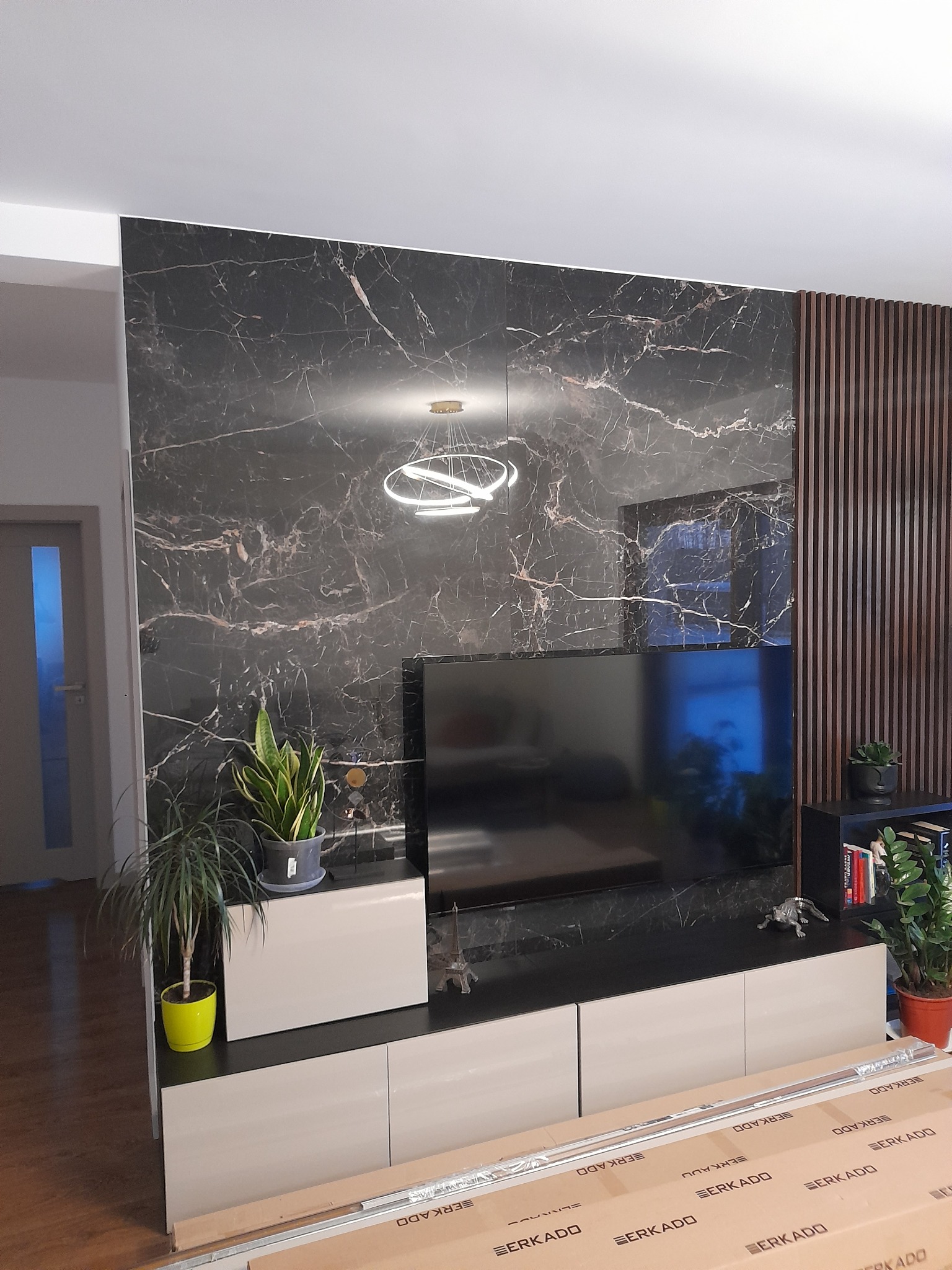 Salon z czarną ścianą imitującą marmur, drewnianymi lamelami, podświetlanym okrągłym żyrandolem i niską szafką pod telewizorem z roślinami doniczkowymi.