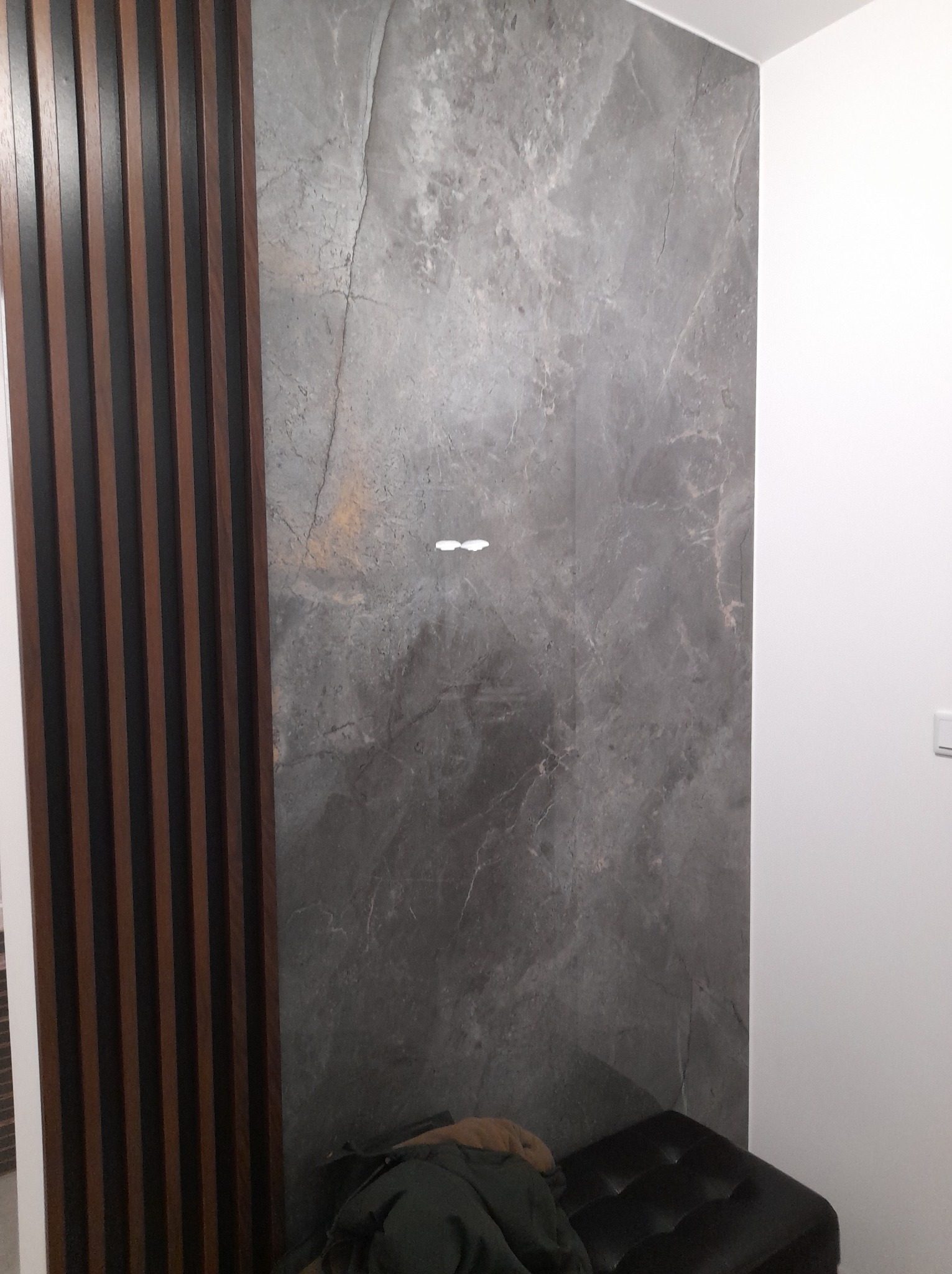 Szara ściana wykończona płytami imitującymi marmur, sąsiadująca z drewnianą ścianką działową z ciemnymi listwami i białą ścianą, z fragmentem czarnego pufa w dolnej części kadru.