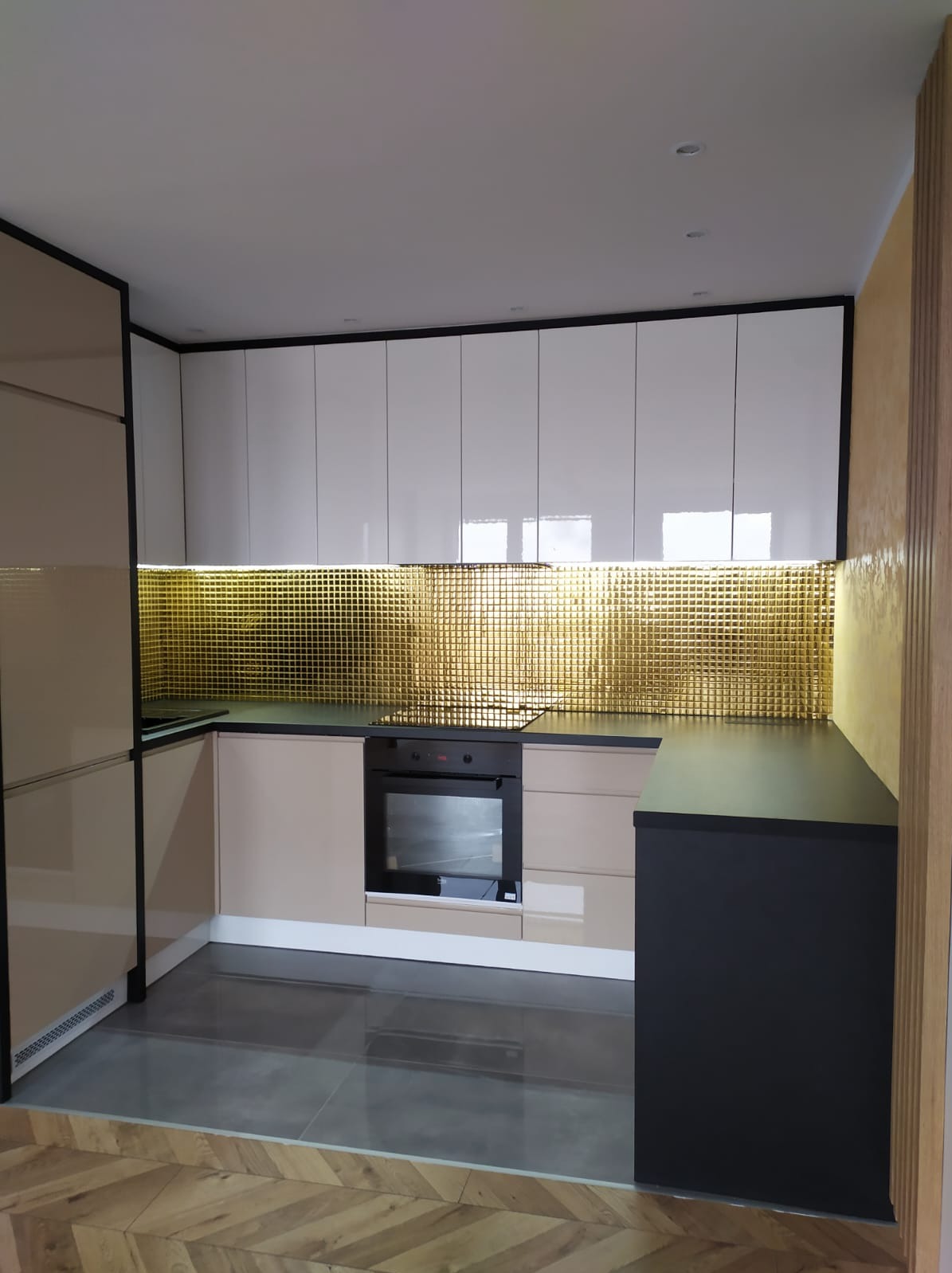 Nowoczesna kuchnia z błyszczącymi, białymi szafkami górnymi, beżowymi szafkami dolnymi i złotą mozaiką nad blatem. Czarny blat kontrastuje z jasnymi elementami. Podłoga wyłożona szarymi płytkami...