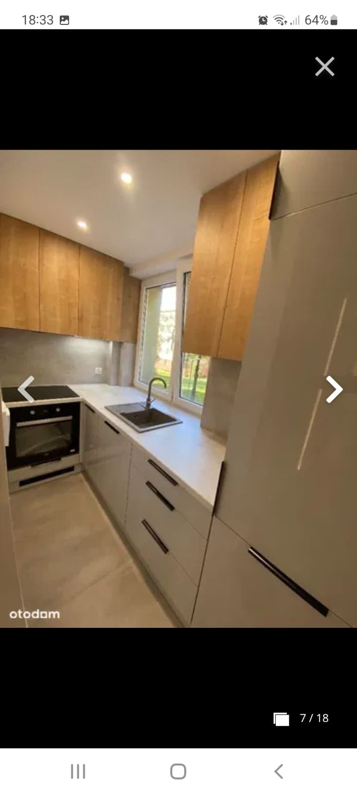 Nowoczesna kuchnia z drewnianymi szafkami wiszącymi i szarymi szafkami dolnymi z czarnymi uchwytami, oświetlona punktowo, z widokiem na zieleń za oknem.