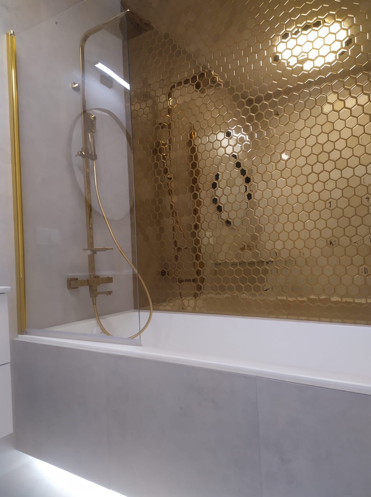 Wykończona łazienka z wanną, złotą armaturą i ścianą z heksagonalnych płytek w odcieniach złota, częściowo zasłonięta szklanym parawanem.