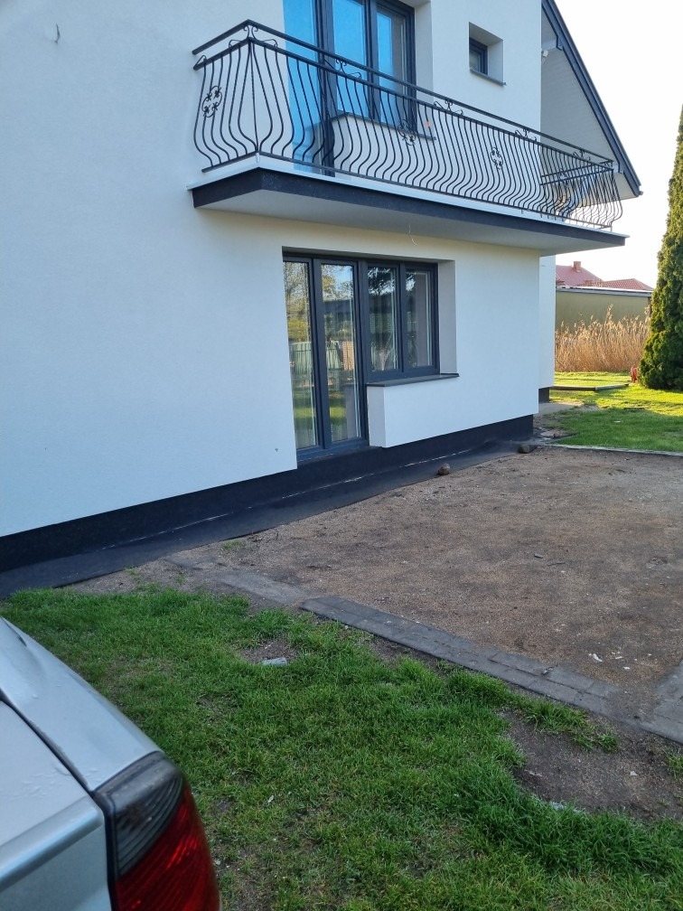 Wykończona elewacja domu jednorodzinnego z balkonem o dekoracyjnej, metalowej balustradzie; widoczne szare okna i cokół.