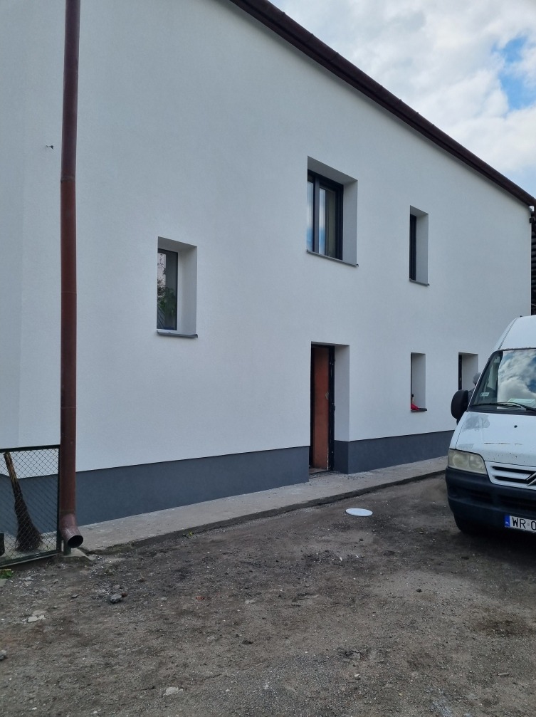 Biała, dwukondygnacyjna elewacja budynku z szarą podmurówką, widoczne okna z czarnymi ramami, brązowa rynna spustowa i zaparkowany biały samochód dostawczy.