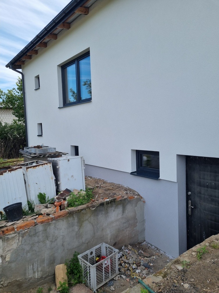 Boczny widok na dom z nową, białą elewacją i szarą podmurówką, widoczne okna z ciemnymi ramami, teren wokół domu częściowo uporządkowany, z pozostałościami po pracach budowlanych.