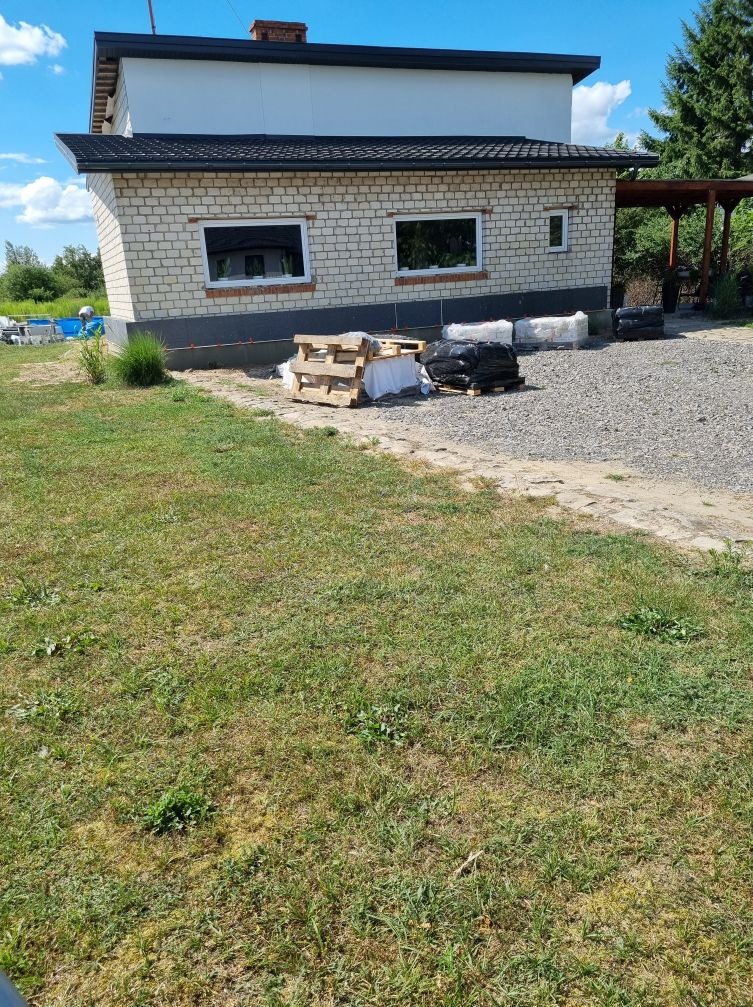 Przygotowania do prac elewacyjnych: ceglany budynek z widocznym ociepleniem fundamentów, paleta i worki z materiałami budowlanymi na żwirowym podłożu.