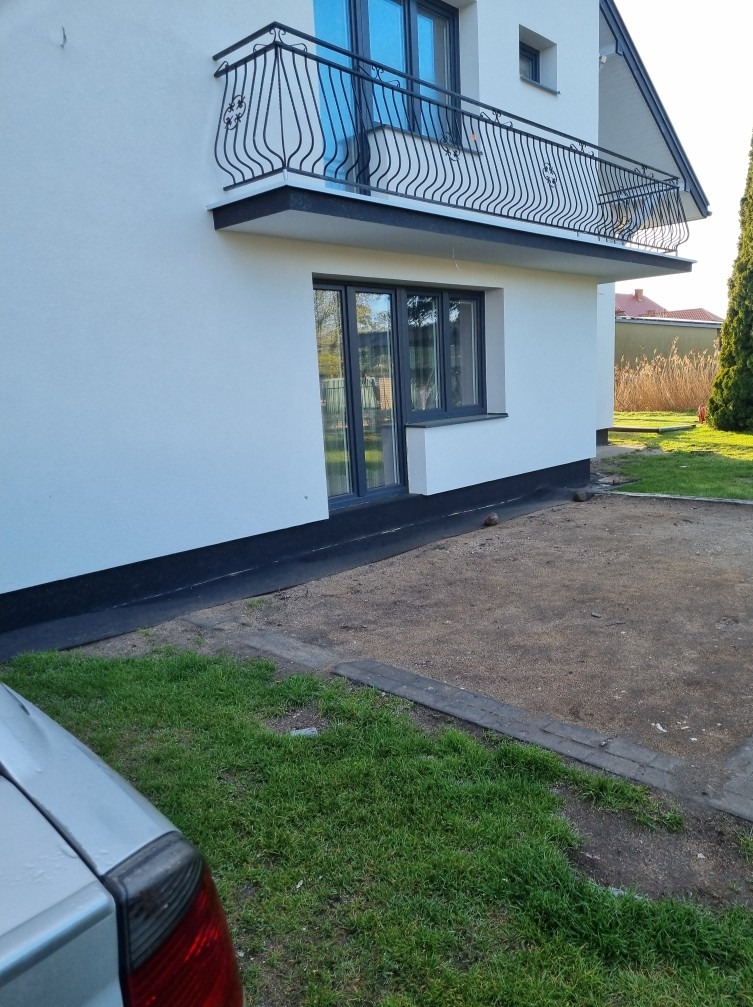 Boczny widok odnowionego domu z białymi ścianami, czarnym wykończeniem, balkonem z ozdobną balustradą i szarymi ramami okiennymi; widoczny fragment samochodu w lewym dolnym rogu.