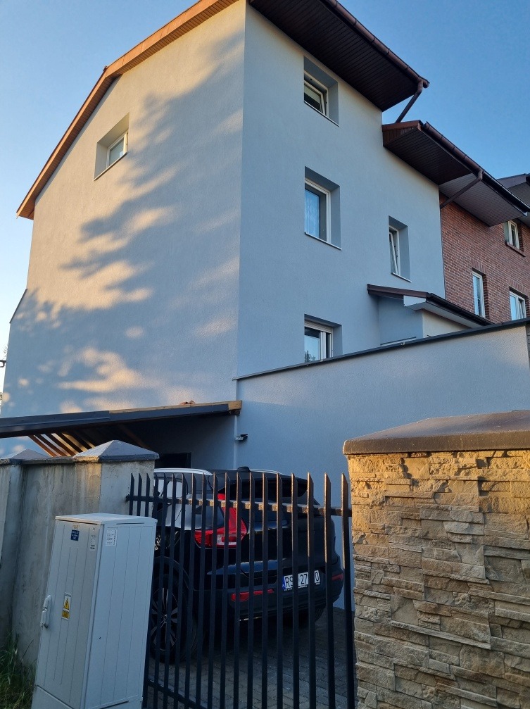 Dwupiętrowy dom z elewacją w odcieniach szarości, widoczny zza metalowego ogrodzenia i kamiennego murku, z zaparkowanym czarnym samochodem pod wiatą.