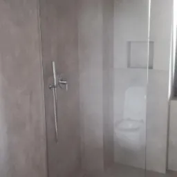 Nowoczesna kabina prysznicowa typu walk-in z szarą armaturą i odpływem liniowym w minimalistycznej łazience wykończonej płytkami w odcieniach szarości, widok na toaletę w tle.