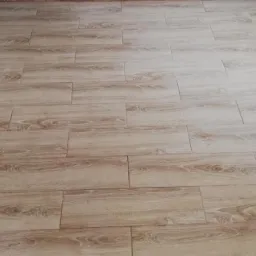 Ułożona podłoga z płytek imitujących drewno w jasnym odcieniu, widoczne fugi i fragment ściany z niedoskonałościami przy podłodze.