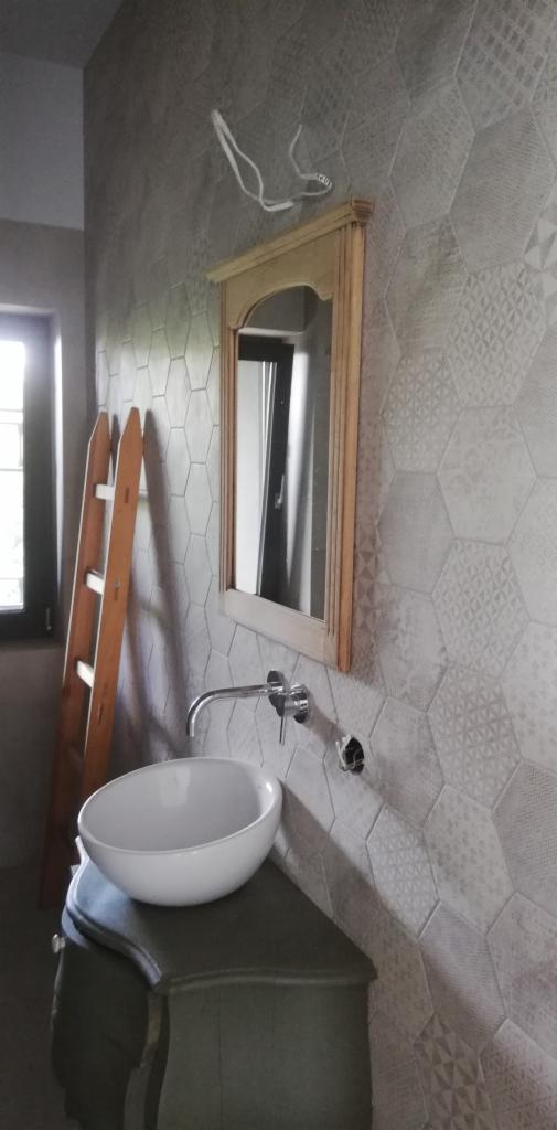 Ukończona łazienka z heksagonalnymi płytkami, umywalką nablatową na szafce w stylu retro, drewnianym lustrem i drabinką opartą o ścianę, widoczne niedokończone podłączenie elektryczne.