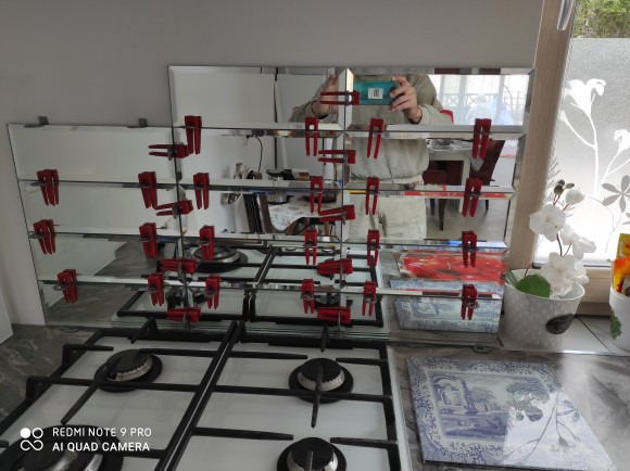 Montaż lustrzanych paneli nad kuchenką gazową, tymczasowo przytrzymywanych czerwonymi klamerkami, widoczne odbicie osoby robiącej zdjęcie telefonem.