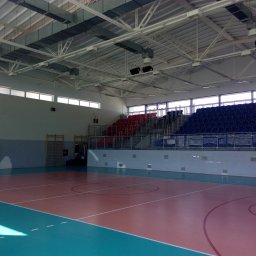 ALTER PROJEKT Sp. z o.o. - Wnętrze nowoczesnej hali sportowej z wielobarwnym parkietem, trybunami w odcieniach czerwieni i błękitu oraz widoczną konstrukcją stalową dachu i systemem wentylacji.
