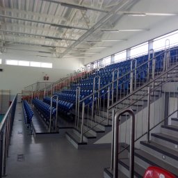 ALTER PROJEKT Sp. z o.o. - Nowoczesna hala sportowa z rzędami niebieskich krzesełek dla widzów, stalowymi balustradami i widocznym fragmentem zielonego boiska. Wysoki sufit z konstrukcją i oświetleniem.