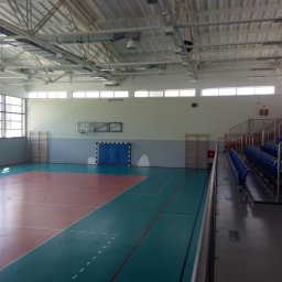 ALTER PROJEKT Sp. z o.o. - Wnętrze hali sportowej z dwukolorową podłogą (czerwień, turkus), koszem, bramką z niebieską siatką i trybunami z pustymi, niebieskimi siedzeniami. Widoczna metalowa konstrukcja dachu.