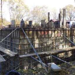 ALTER PROJEKT Sp. z o.o. - Szalunek fundamentów lub ścian piwnicy na budowie, z widoczną gruszką do betonu i pracownikiem na górze konstrukcji, w słoneczny dzień.