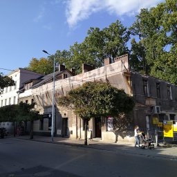 ALTER PROJEKT Sp. z o.o. - Kamienica z szarą elewacją, częściowo zakryta siatką remontową, z widocznymi kominami i żurawiem budowlanym, obok białego budynku i ulicy.