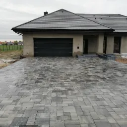 Nowoczesny podjazd z szarej kostki brukowej o nieregularnym wzorze, prowadzący do domu z garażem i numerem 7, w tle widoczne inne domy.