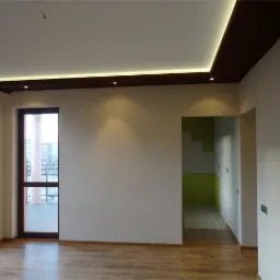 Pusty pokój z drewnianą podłogą, białymi ścianami i podwieszanym sufitem z oświetleniem LED, widok na balkon przez ciemne, drewniane drzwi balkonowe oraz zielone schody w tle