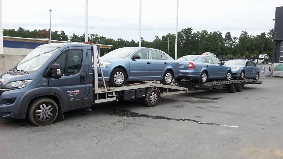 Laweta z trzema niebieskimi samochodami marki Skoda Octavia, zaparkowana na szarym asfalcie przed budynkiem, widoczny napis 'TRANSCAR' na pojeździe.