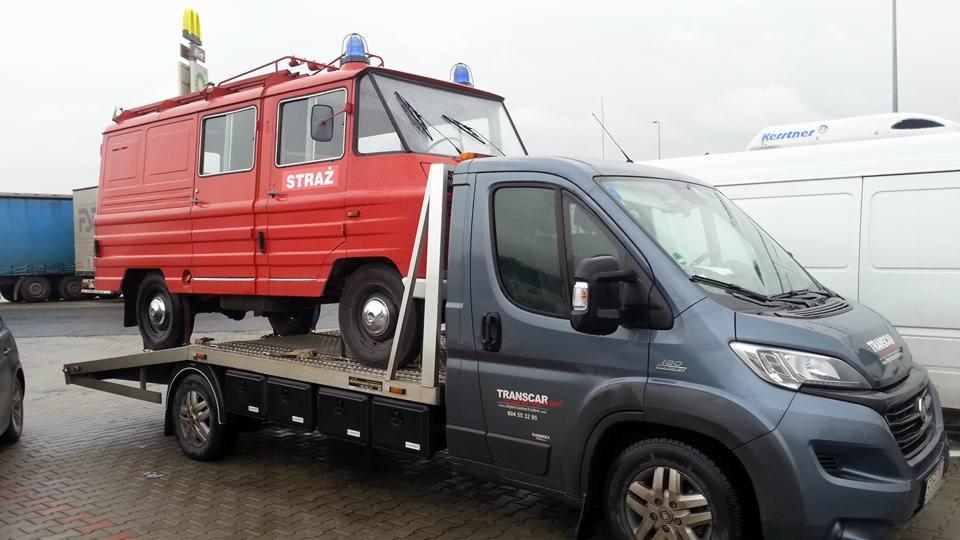 Czerwony, zabytkowy wóz strażacki z niebieskimi kogutami przewożony na platformie lawety marki Fiat Ducato z logo Transcar na tle parkingu.