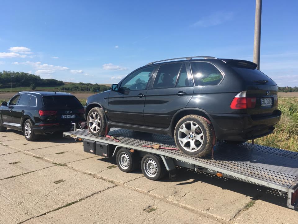 Czarny SUV BMW X5 na perforowanej platformie lawety, za nim czarny Audi Q7, oba auta na betonowej nawierzchni, w tle pole i błękitne niebo.