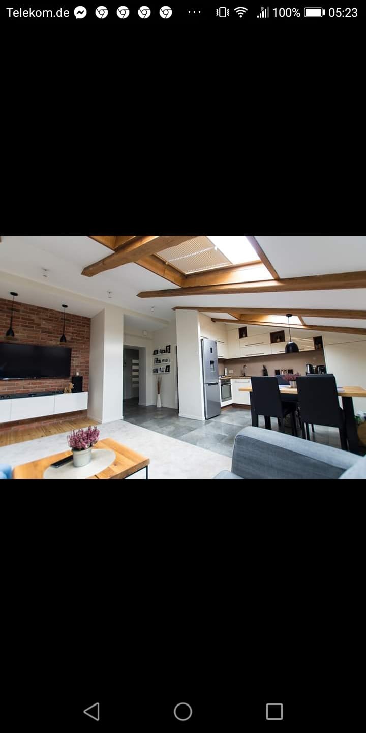 Przestronne wnętrze mieszkania z drewnianymi belkami stropowymi, ceglaną ścianą z telewizorem, aneksem kuchennym i stołem z krzesłami.