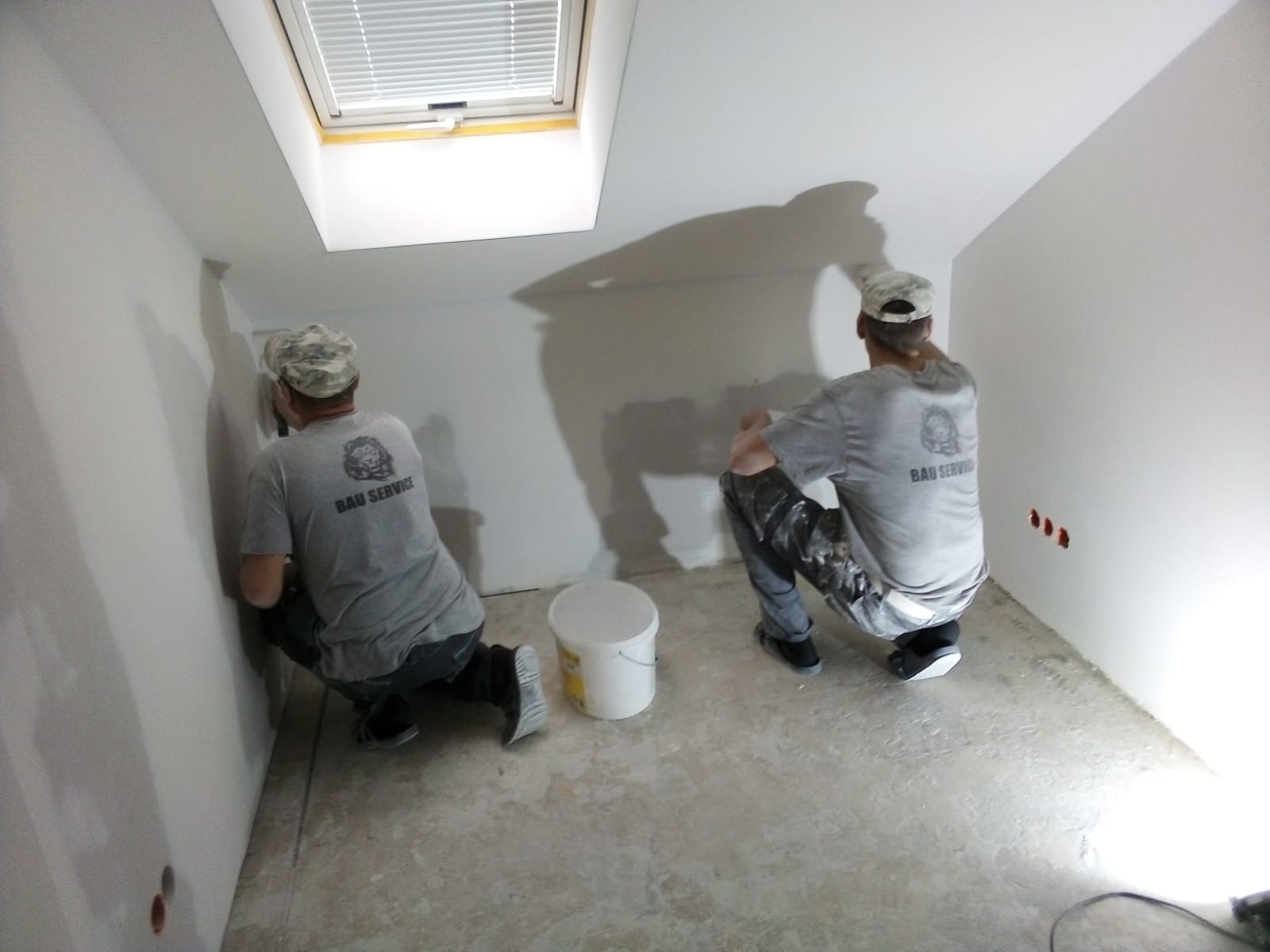Dwóch mężczyzn w koszulkach z logo firmy wygładza szpachelką ściany na poddaszu z oknem dachowym, widoczne puszki elektryczne i wiadro z masą szpachlową.